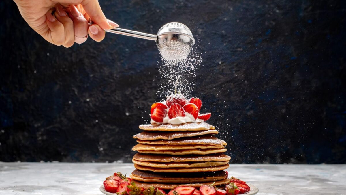 a front view of yummy and round pancakes with cream, powdered sugar, and red strawberries